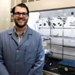 Spencer wears a lab coat and stands in front of a series of test tubes.