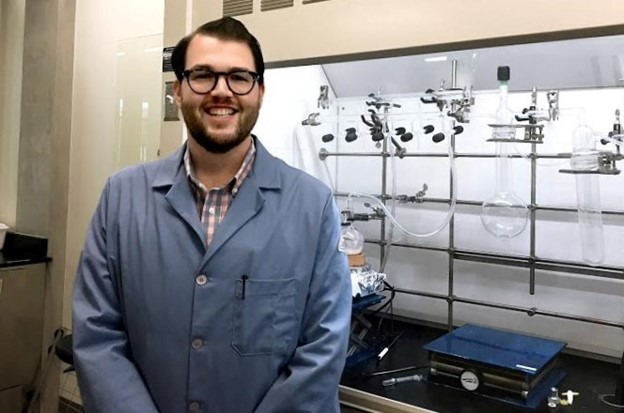 Spencer wears a lab coat and stands in front of a series of test tubes.