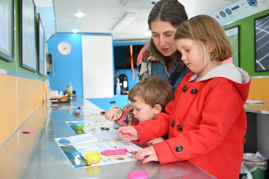 An adult helps two children complete an activity with playdoh in the mobile classroom.