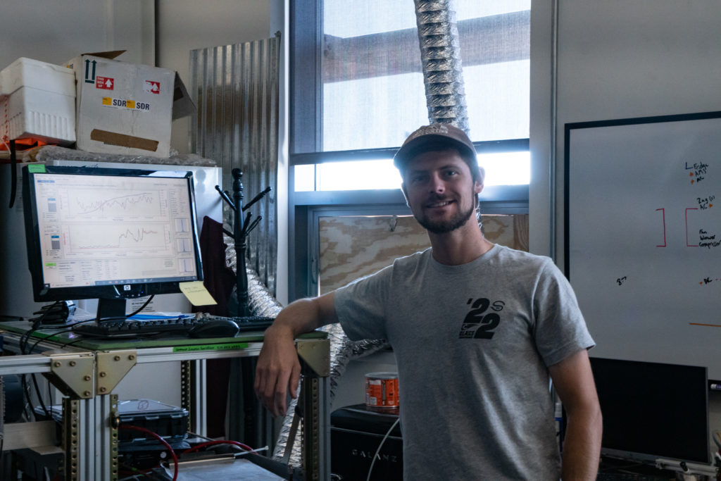 A man stands next to a computer in a workshop, smiling confidently with equipment and materials in the background.