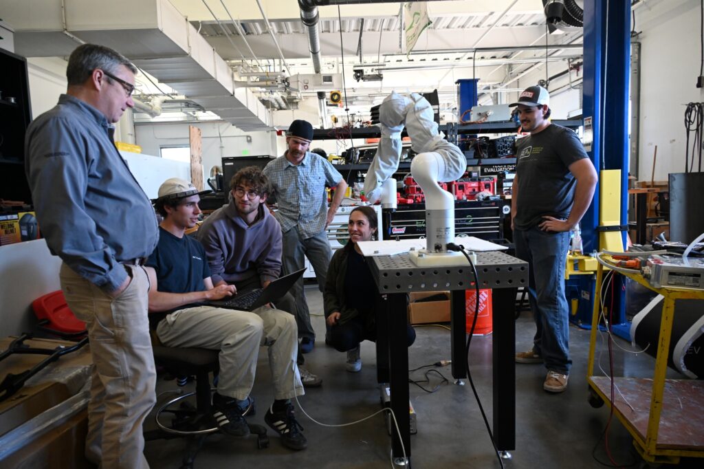 5 students stand around a robotic arm on a table with their advisor looking on.