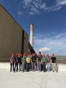 A large group stands on a roof in front of a coal plant smoke stack